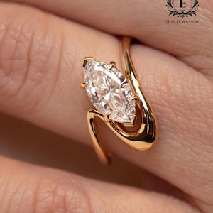 May include: A marquise-cut diamond ring with a unique, flowing gold band. The diamond is set in a four-prong setting, and the band curves around the finger. The ring is a close-up shot, highlighting the details of the jewelry.