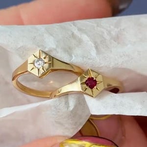 May include: Two gold rings with a starburst design. One ring features a clear gemstone, the other a red gemstone. The rings are displayed on white tissue paper. The rings are from EliteJewelsArt.
