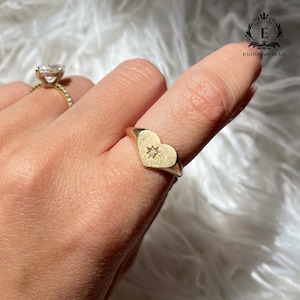 May include: A gold ring with a heart-shaped face, featuring a star design and a small diamond. The ring is worn on a finger. Another gold ring with a large diamond is visible in the background.