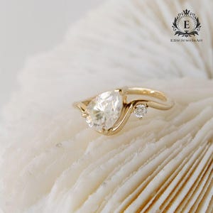 Puede incluir: Un anillo de compromiso de oro con un diamante en forma de pera y un pequeño diamante de acento. El anillo está sobre un fondo blanco.