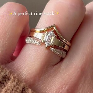 May include: A stack of three gold rings. The top ring has a chevron design. The middle ring features a large, emerald-cut diamond. The bottom ring is encrusted with small diamonds. The text "A perfect ring stack" is visible.