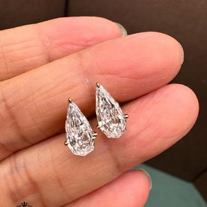 May include: A pair of pear-shaped clear gemstones set in gold metal prongs. Each stone features a faceted cut that reflects light, creating a brilliant appearance. The earrings are held in place, showcasing the pointed top and rounded base design.