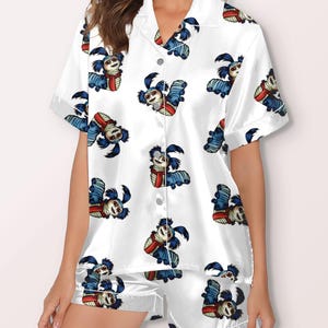 Op de afbeelding: Witte satijnen pyjamaset met een herhalend patroon van een stripfiguur met blauwe vacht, een rode sjaal en een lachend gezicht. De set bevat een shirt met korte mouwen en bijpassende shorts.