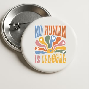 Può includere: Un bottone bianco con la scritta "NO HUMAN IS ILLEGAL" in stile retrò. Il design include un fiore colorato con petali a forma di cuore nei toni dell'arancione, giallo, verde e blu. Il bottone ha un retro con spilla metallica.