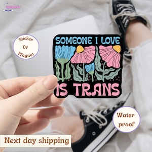 May include: A black sticker with the text "SOMEONE I LOVE IS TRANS" in white and pink. The sticker features blue and pink floral illustrations with green stems and leaves. The sticker is held by a hand.