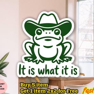 May include: A green frog wearing a cowboy hat sticker with the phrase "It is what it is". The sticker is waterproof and can be used as a magnet. The design is a cartoon style.