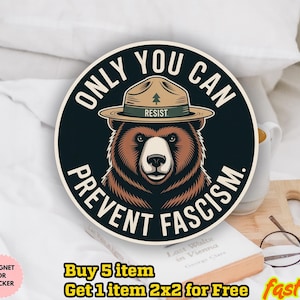 May include: A round sticker with a bear wearing a ranger hat. The sticker has the text "ONLY YOU CAN PREVENT FASCISM" in a circular design. The bear is brown and tan, and the hat is tan. The word "RESIST" is on the hat.