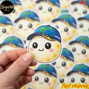 Puede incluir: Una pegatina con una luna de dibujos animados sonriente que lleva una gorra de béisbol azul con un diseño de globo terráqueo. La luna tiene mejillas rosadas y cráteres amarillos. La palabra "ARTEMIS" está en la gorra. Múltiples pegatinas del mismo diseño en el fondo.