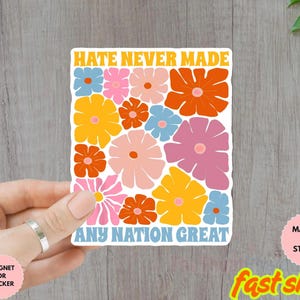 Puede incluir: Una pegatina colorida con el texto "HATE NEVER MADE ANY NATION GREAT" en naranja y azul. La pegatina presenta un diseño floral con flores naranjas, rosas, amarillas y azules. La pegatina está sujeta por una mano.