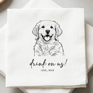 May include: White linen napkins with a black line drawing of a Golden Retriever dog and the text "drink on us!" in a script font. The napkins are stacked on a light gray surface, with a small plate and a sprig of lavender in the background.