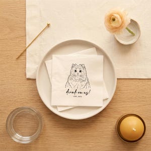 May include: A white square napkin with a black line drawing of a cat's face and the text "drink on us!" is on a white plate. A gold cocktail pick, a small bowl with a flower, a candle, and a glass are also on the wooden table.