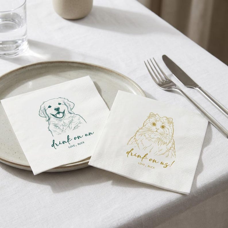 May include: Two white paper napkins with illustrations of a dog and a cat. The dog is green and the cat is gold. Each napkin has the phrase "Drink on us!" and "Love, Buck" printed on it. A fork and knife are on the right.