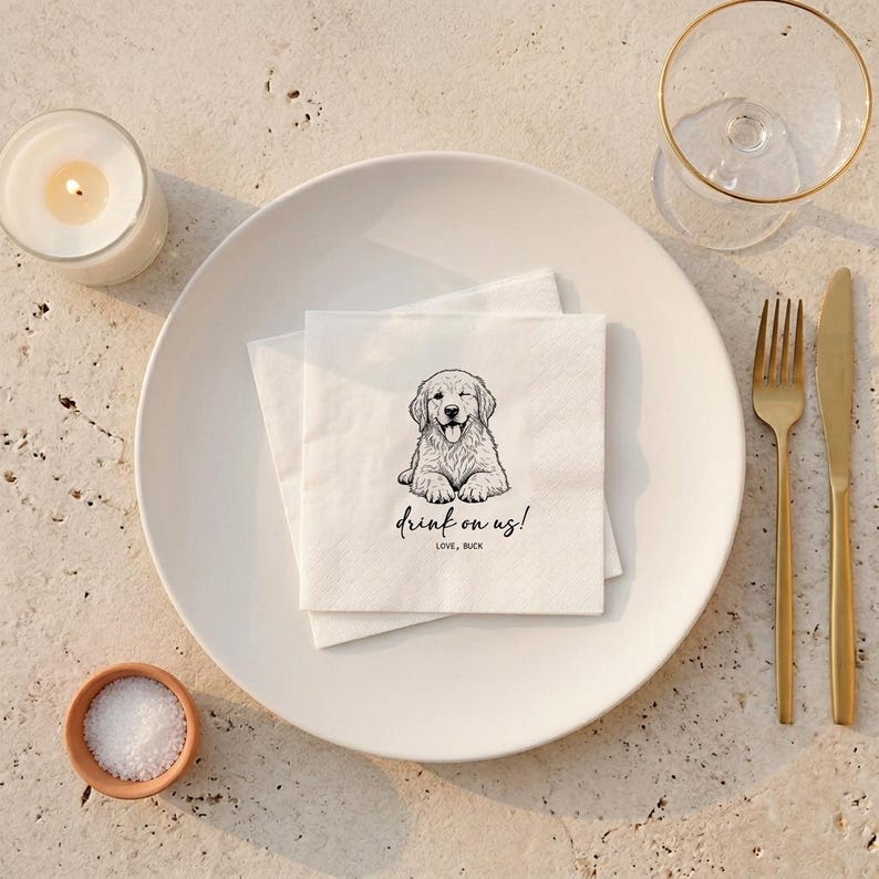 May include: White paper napkins with a black and white illustration of a Golden Retriever puppy winking and sticking out its tongue. The napkins have the text "drink on us!" and "LOVE, BUCK". The napkins are on a white plate with gold silverware.