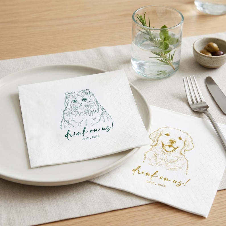 May include: White paper napkins with pet illustrations. One napkin features a cat with the text "drink on us!" in green. The other napkin has a dog with the same text in gold. A glass of water with herbs and a small bowl of olives are also present.