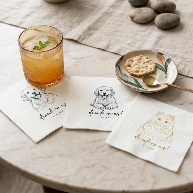 May include: A refreshing drink in a textured glass with ice and mint, alongside three white napkins. Each napkin features a different pet illustration with the text "drink on us!" and "LOVE, BUCK". A small plate with a cracker and spoon is also present.