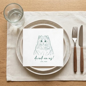 May include: White square paper napkin with a green illustration of a cat's face and the text "drink on us!" and "LOVE, BUCK". The napkin is placed on a white plate with a fork and knife. A glass of water is to the left.