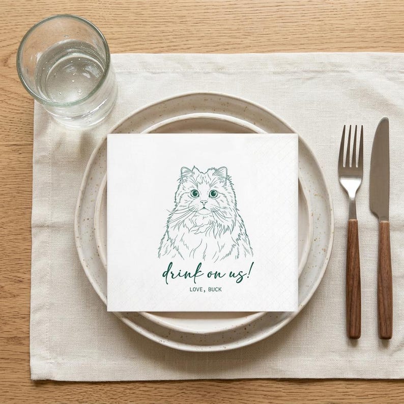May include: White square paper napkin with a green illustration of a cat's face and the text "drink on us!" and "LOVE, BUCK". The napkin is placed on a white plate with a fork and knife. A glass of water is to the left.