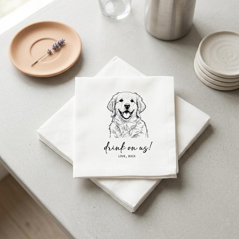 May include: White linen napkins with a black line drawing of a Golden Retriever dog and the text "drink on us!" in a script font. The napkins are stacked on a light gray surface, with a small plate and a sprig of lavender in the background.