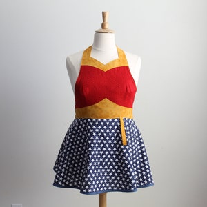 May include: A red and yellow Wonder Woman-themed apron with a blue and white star pattern. The apron has a halter neck strap and a gathered skirt.