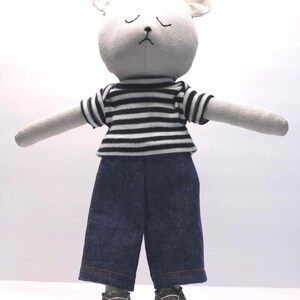 May include: A handmade stuffed animal in the shape of a bear with a white head and ears, wearing a black and white striped shirt, blue denim pants, and gray sneakers. The bear has closed eyes and outstretched arms.