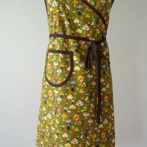 May include: A green wrap apron with a brown trim and a pocket. The apron is covered in a mushroom print with red, yellow, and white mushrooms.