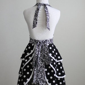 May include: A black and white polka dot apron with a halter neck and tiered skirt design. The apron features a contrasting floral pattern on the neck tie and waist tie. The apron is displayed on a mannequin.