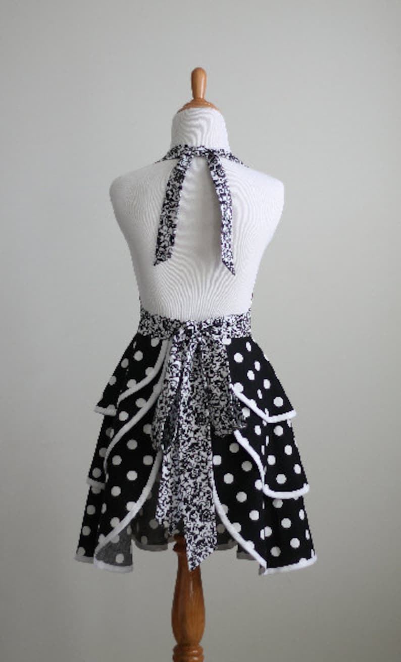 May include: A black and white polka dot apron with a halter neck and tiered skirt design. The apron features a contrasting floral pattern on the neck tie and waist tie. The apron is displayed on a mannequin.