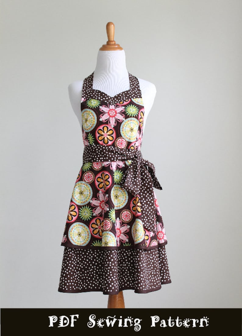 May include: A brown apron with a floral pattern and white polka dots, displayed on a mannequin. The apron features a halter neck, a waist tie, and a tiered skirt. The floral design includes pink, yellow, and green elements. The text "PDF Sewing Pattern" is at the bottom.