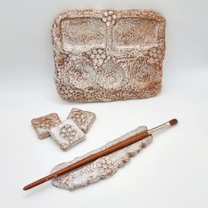 May include: A white ceramic paint palette with four wells and a brush holder. The palette has a textured surface and is decorated with a brown glaze. Three small square ceramic dishes are also included.