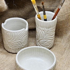 May include: A set of three off-white ceramic art studio containers. The set includes a small bowl, a cylindrical brush holder with leaf design, and a small cup with a leaf design. The brush holder contains three paint brushes.