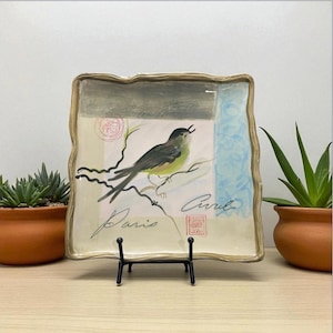May include: Square decorative plate with a bird illustration. The plate features a bird perched on a branch, with the word "Paris" written in script. The plate has a wavy, gold-coloured border and is displayed on a black metal stand.