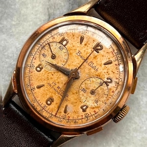 Peut inclure: Montre Brevets vintage dorée avec un bracelet en cuir marron. Le cadran de la montre est de couleur crème avec des chiffres noirs et des sous-cadrans. La montre possède une fonction chronographe et le texte "17 RUES ANTIMAGNETIC" est visible.
