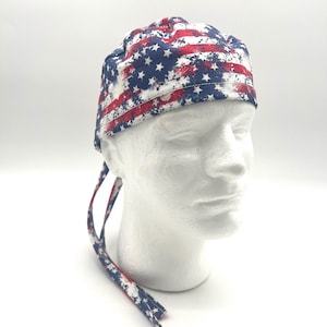 May include: A scrub cap featuring a distressed American flag pattern with red, white, and blue colors. The cap has a rounded crown and two long fabric ties extending from the back, designed to be secured around the head for a comfortable fit.