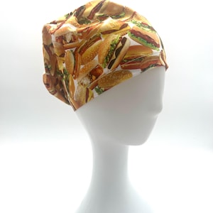 May include: A scrub cap featuring a repeating pattern of various cheeseburgers and hamburgers on a white background. The fabric displays detailed illustrations of buns, lettuce, cheese, and meat patties in shades of brown, green, and yellow.