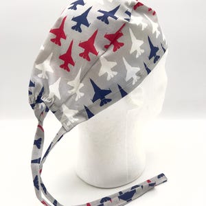 May include: Gray surgical scrub hat with a repeating pattern of red, white, and blue fighter jets. The hat has two long ties that are tied at the back of the head.