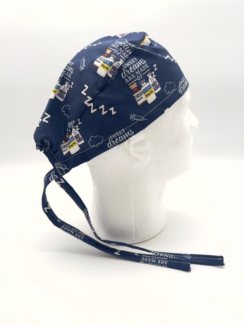 Op de afbeelding: Blauwe chirurgische scrub-muts met een witte print van medicijnflesjes, spuiten en de tekst "Sweet dreams are made of ZZZ".