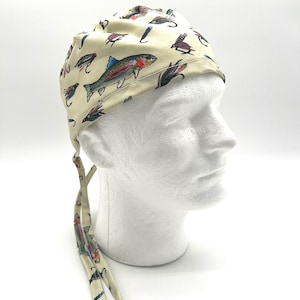 May include: A cream-colored fishing cap featuring a repeating pattern of colorful fish and fishing lures. The cap has a tie closure at the back and is designed to be worn on the head. The design includes a rainbow trout and various fly fishing lures.