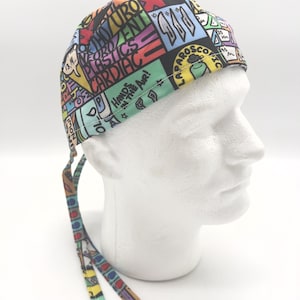 May include: A colorful surgical scrub hat with a black background and a variety of medical terms printed on it, including "Hands in the Air!" and "Laparoscopic". The hat has two long ties.