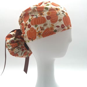 May include: A patterned scrub cap featuring orange pumpkins and fall floral designs on a cream background. The cap has a brown ribbon and is designed to be worn with a ponytail. Perfect for healthcare professionals.