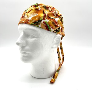 May include: A patterned surgical scrub cap featuring a repeating design of hamburgers, sandwiches, and other fast food items. The cap is tied in the back with matching fabric ties. The background is white.