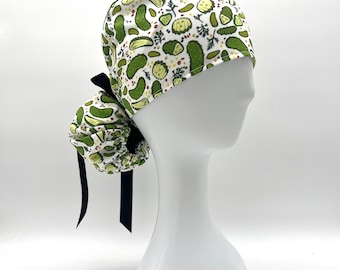 May include: A white and green scrub cap featuring a pattern of green pickles and yellow flowers. The cap has a black bow and is displayed on a white mannequin head. The cap is designed for medical professionals.