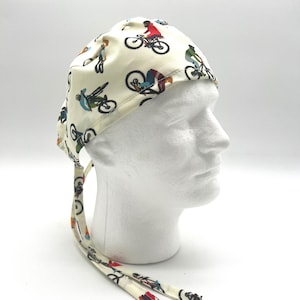 May include: Cream-colored surgical scrub cap featuring a repeating pattern of cyclists in various poses and colorful outfits. The cap has ties for a secure fit. Ideal for medical professionals or anyone who enjoys cycling.