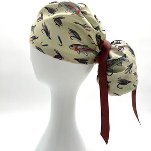 May include: A beige scrub cap featuring a repeating pattern of colorful trout and fishing flies. The cap is tied with a dark red ribbon. The cap is designed to be worn on the head and is shown on a white mannequin head.