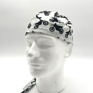 May include: A white head wrap featuring a repeating pattern of black motorcycles. The head wrap is tied at the back and is displayed on a white mannequin head. The fabric has a light, slightly textured appearance.