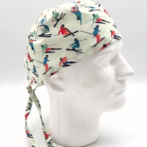 May include: A white scrub hat with a colorful pattern of skiers on a white background. The hat has two long ties that are tied at the back of the head.