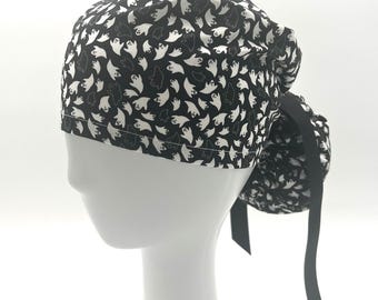 May include: A black and white patterned scrub cap featuring a repeating ghost design. The cap is tied in the back with a black ribbon. The cap is displayed on a white mannequin head.
