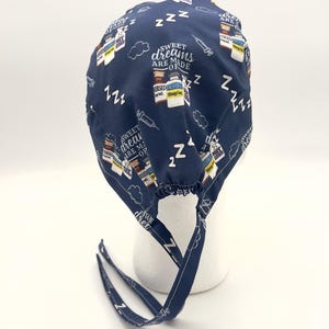 Op de afbeelding: Een donkerblauwe chirurgische scrub-muts met een witte print van medicijnflesjes, spuiten en de tekst "Sweet dreams are made of ZZZ". De muts heeft twee lange banden.