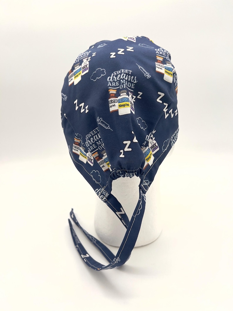 Op de afbeelding: Een donkerblauwe chirurgische scrub-muts met een witte print van medicijnflesjes, spuiten en de tekst "Sweet dreams are made of ZZZ". De muts heeft twee lange banden.