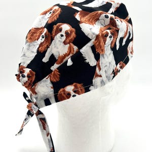 May include: Black scrub hat with a repeating pattern of Cavalier King Charles Spaniel dogs. The hat has a tie on the back.