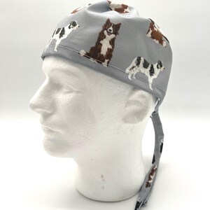 May include: A light gray surgical scrub cap featuring a repeating pattern of various dog breeds in brown, black, and white. The cap is tied at the back with matching fabric ties. The dogs are in different poses.